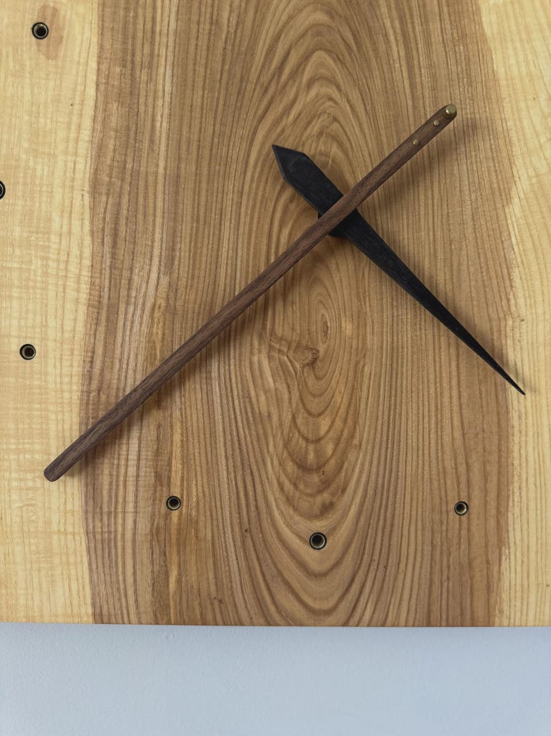 Pu&ograve; includere: Un orologio da parete in legno con finitura a venatura naturale. L'orologio presenta una lancetta delle ore marrone scuro e una lancetta dei minuti nera. Il quadrante dell'orologio ha piccoli indicatori circolari neri.