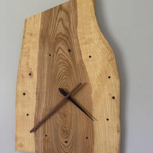 Pu&ograve; includere: Un orologio da parete in legno dalla forma irregolare e naturale. Il quadrante &egrave; in legno chiaro con venature scure. Presenta indicatori delle ore neri e lancette marroni scure. L'orologio &egrave; montato su una parete grigio chiaro.