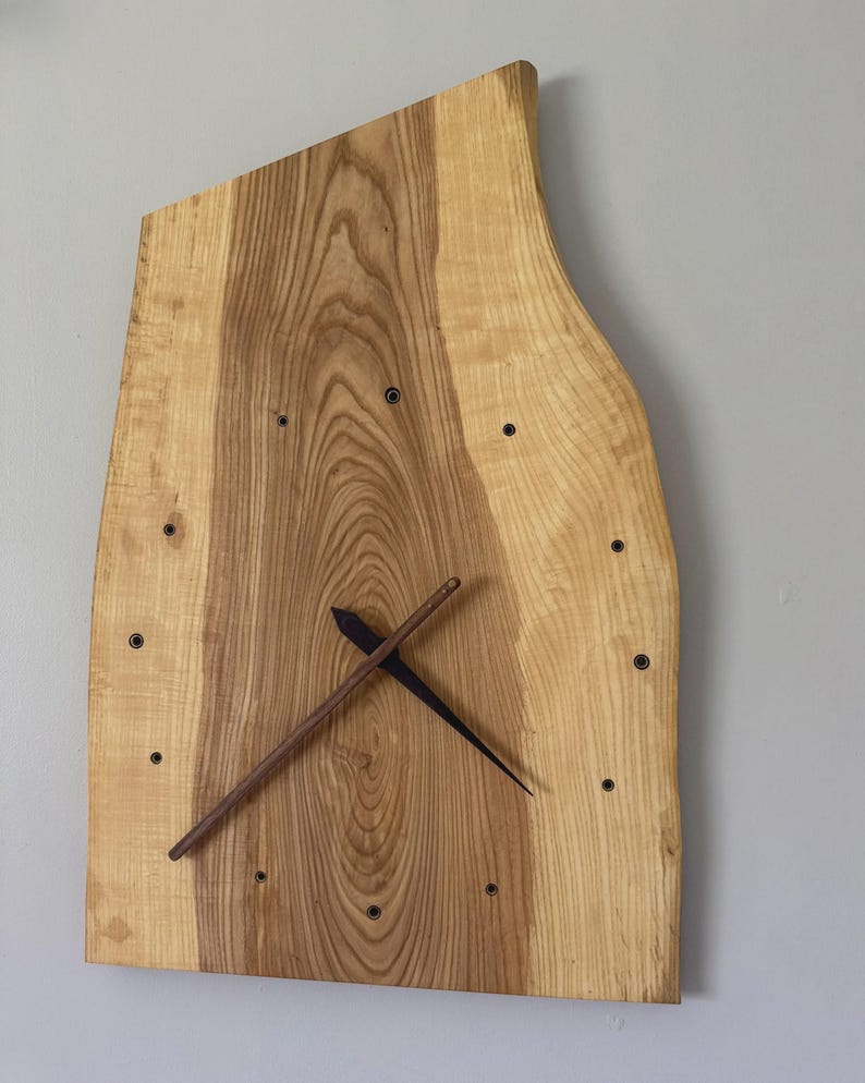 Pu&ograve; includere: Un orologio da parete in legno dalla forma irregolare e naturale. Il quadrante &egrave; in legno chiaro con venature scure. Presenta indicatori delle ore neri e lancette marroni scure. L'orologio &egrave; montato su una parete grigio chiaro.