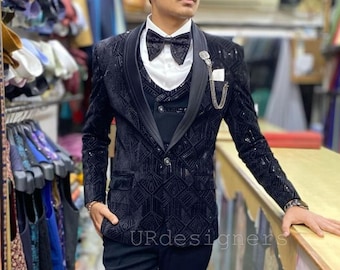 May include: A black velvet suit with a patterned design. The suit includes a jacket, vest, and pants. The jacket has a shawl collar and a single button closure. The vest has a V-neck and a button closure. The pants have a straight leg. The model is wearing a black bow tie and sunglasses.