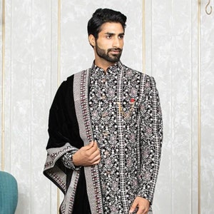 May include: A black velvet shawl with a silver and red embroidered border. The shawl is draped over a man wearing a black and silver embroidered kurta and white pants. He is wearing brown leather shoes.