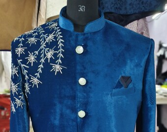 Può includere: Una giacca in velluto blu turchese con ricami argentati sulle maniche e una tasca sul lato sinistro. La giacca ha un colletto alla coreana e bottoni bianchi. Il testo "Urdesigners" è visibile sulla giacca.