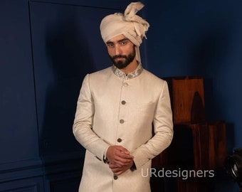 Puede incluir: Un hombre con una kurta blanca de manga larga con un cuello bordado en plata, pantalones blancos y mocasines marrones. Lleva un turbante blanco y tiene barba. El fondo es una pared azul oscuro con un estante de madera.