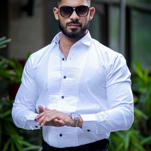 May include: A white dress shirt with a pleated front and black buttons. The shirt has long sleeves and is paired with black pants. The image also shows sunglasses and a wristwatch.