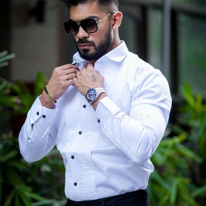 May include: A white dress shirt with black buttons and a pleated front. The shirt is paired with black pants. The person is wearing a watch, sunglasses, and a bracelet.