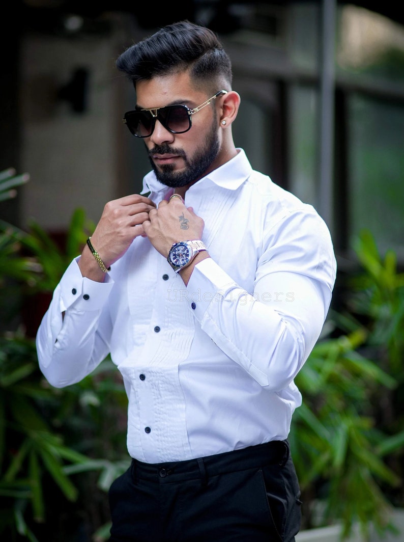 May include: A white dress shirt with black buttons and a pleated front. The shirt is paired with black pants. The person is wearing a watch, sunglasses, and a bracelet.