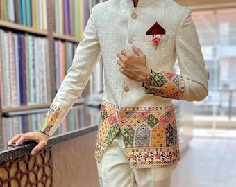 May include: A white blazer with a mandarin collar and gold buttons. The blazer has a colorful patterned trim around the bottom and cuffs. The blazer is worn over a white shirt and white pants.