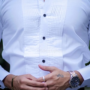 May include: White dress shirt with a pleated bib and black buttons. The shirt has a classic design, suitable for formal occasions. The sleeves are long with buttoned cuffs.