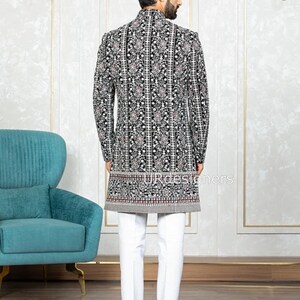 May include: A black velvet long-sleeved jacket with a white and silver floral pattern. The jacket has a mandarin collar and a button closure. The model is wearing white pants and brown shoes.