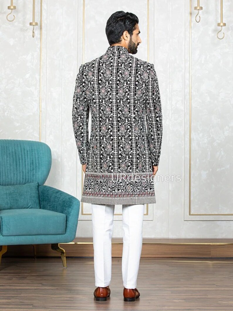 May include: A black velvet long-sleeved jacket with a white and silver floral pattern. The jacket has a mandarin collar and a button closure. The model is wearing white pants and brown shoes.