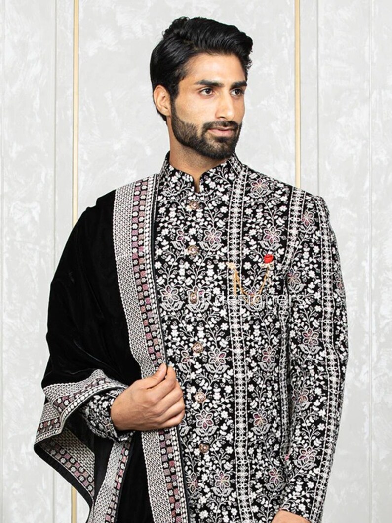 May include: A black velvet shawl with a silver and pink embroidered border. The shawl is draped over a man wearing a black embroidered jacket with a white floral pattern.