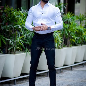 May include: A man wearing a white dress shirt with a pleated front and black pants. The shirt has black buttons and is paired with black dress pants and black boots. The man is also wearing sunglasses and a watch.