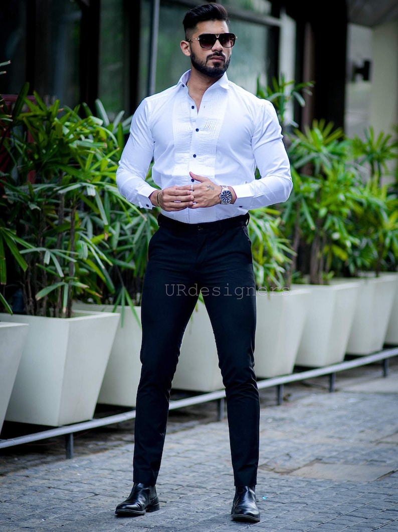 May include: A man wearing a white dress shirt with a pleated front and black pants. The shirt has black buttons and is paired with black dress pants and black boots. The man is also wearing sunglasses and a watch.