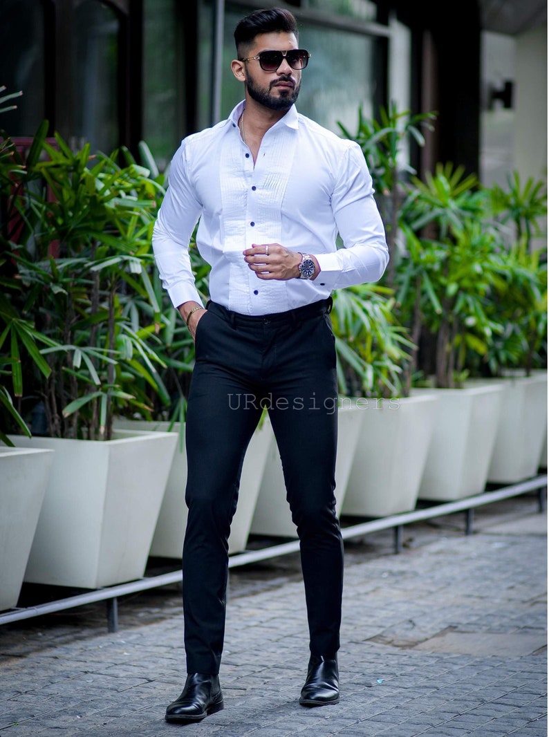 May include: A white dress shirt with a pleated front and black buttons, paired with black trousers and black boots. The image also shows a watch and sunglasses. The text "URdesigners" is visible.