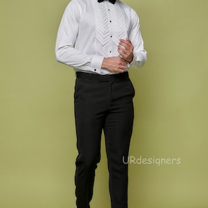 May include: A white dress shirt with a pleated front and a black bow tie. The shirt has long sleeves and a button-up collar. The man is wearing black pants and black shoes.