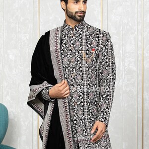 May include: A black embroidered kurta with a white and silver floral pattern. The kurta has a mandarin collar and long sleeves. The man is wearing a black velvet shawl with a silver border.