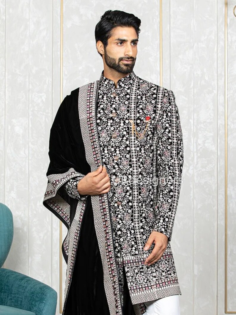 May include: A black embroidered kurta with a white and silver floral pattern. The kurta has a mandarin collar and long sleeves. The man is wearing a black velvet shawl with a silver border.