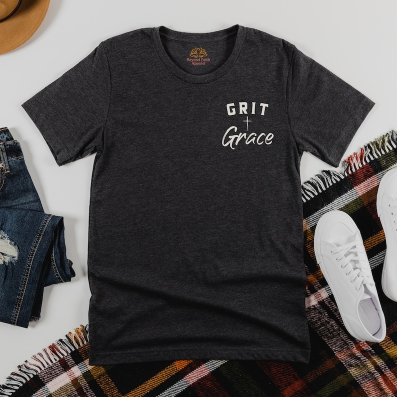Op de afbeelding: Een donkergrijs T-shirt met de tekst "GRIT + Grace" erop gedrukt. Het shirt ligt plat op een geruite deken met een paar witte sneakers en een blauwe spijkerbroek.