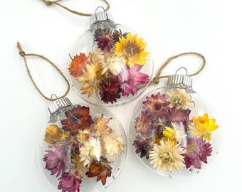 May include: Three clear glass ornaments filled with dried flowers. The ornaments are decorated with a variety of dried flowers in shades of pink, yellow, and orange. The ornaments are hanging from a white background.