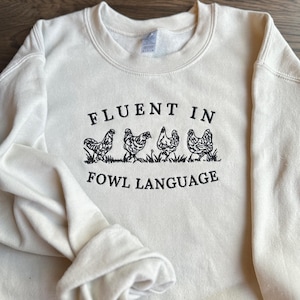 May include: A cream-colored sweatshirt with the text "Fluent in Fowl Language" and a line of five black chickens walking across the front.