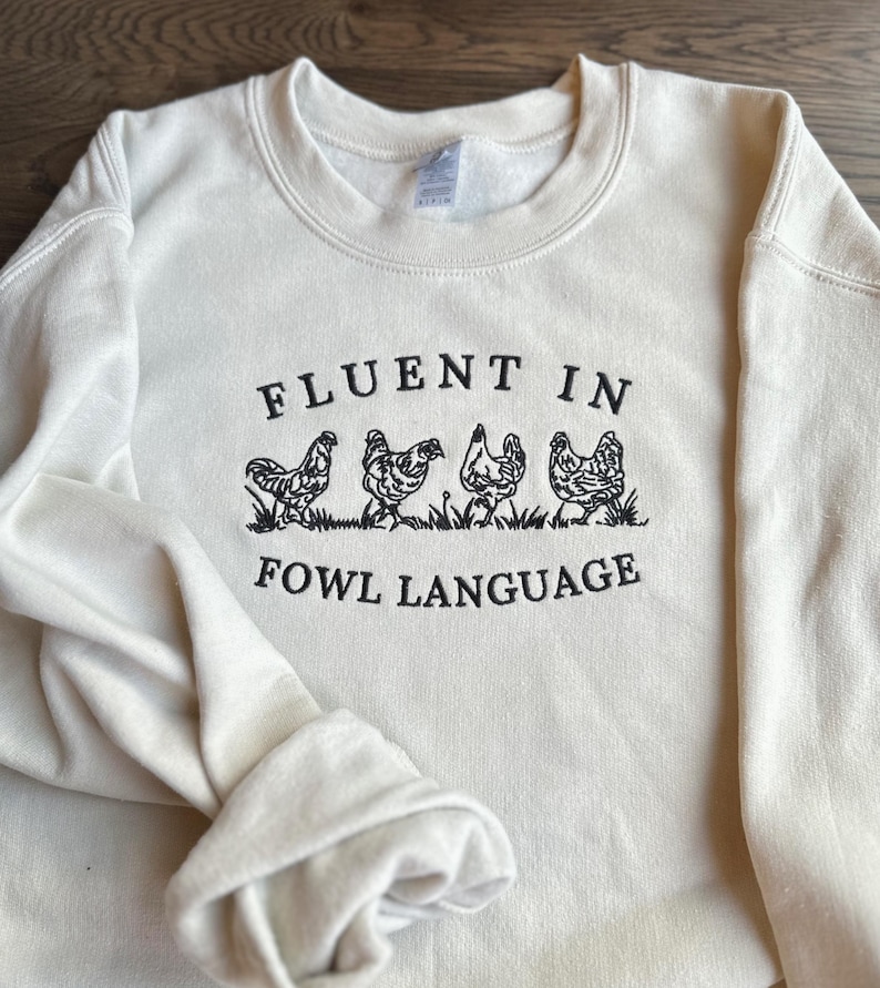 May include: A cream-colored sweatshirt with the text "Fluent in Fowl Language" and a line of five black chickens walking across the front.