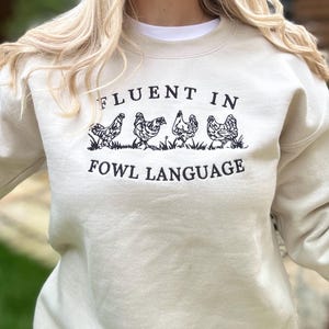 May include: A light beige sweatshirt with the text "Fluent in Fowl Language" and a graphic of four chickens walking in a line.
