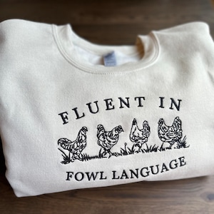 May include: A cream-colored sweatshirt with black embroidered text that reads "Fluent in Fowl Language".  The text is surrounded by five black embroidered chickens.