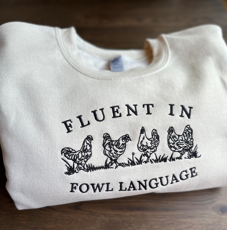 May include: A cream-colored sweatshirt with black embroidered text that reads "Fluent in Fowl Language".  The text is surrounded by five black embroidered chickens.