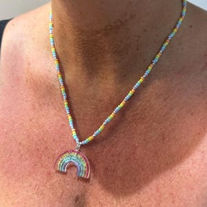 May include: A necklace featuring a strand of small, multicolored beads in shades of yellow, pink, blue, and green, interspersed with silver-toned accents. A translucent, arched rainbow pendant with a striped pattern hangs from the center.