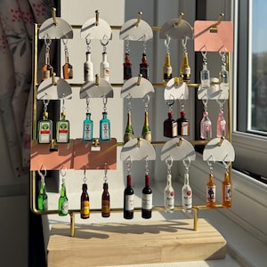 Op de afbeelding: Een display van miniatuur sleutelhangers in de vorm van drankflessen. De sleutelhangers zijn gerangschikt op een goudkleurig metalen rek met witte en roze accenten. De flessen hebben verschillende kleuren en vormen, met zilveren sleutelringen.