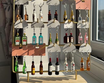 May include: A display of miniature liquor bottle keychains. The keychains are arranged on a gold-colored metal rack with white and pink accents. The bottles are various colors and shapes, including brown, green, and clear, with silver key rings.