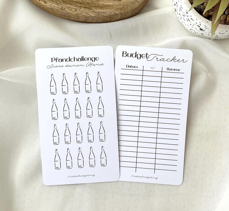 May include: Two white cards with black text and illustrations. One card says "Pfandchallenge" and features bottle drawings. The other card is a "Budget Tracker" with columns for date, +/- and sum. Both cards have the text "fridasbudgeting."