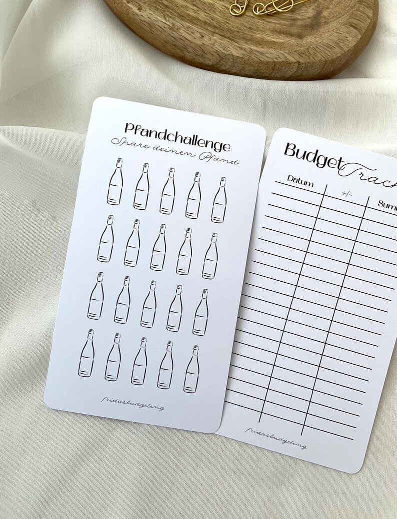 May include: Two white cards with black text and illustrations. One card has a bottle illustration and the text "Pfandchallenge Spare deinen Pfand". The other card has a budget tracker with the text "Budget Tracker".