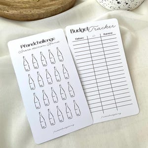 May include: Two white cards with black text and illustrations. One card reads "Pfandchallenge Spare deinen Pfand" with bottle illustrations. The other card says "Budget Tracker" with columns for tracking expenses. Both cards have the text "fridasbudgetling".