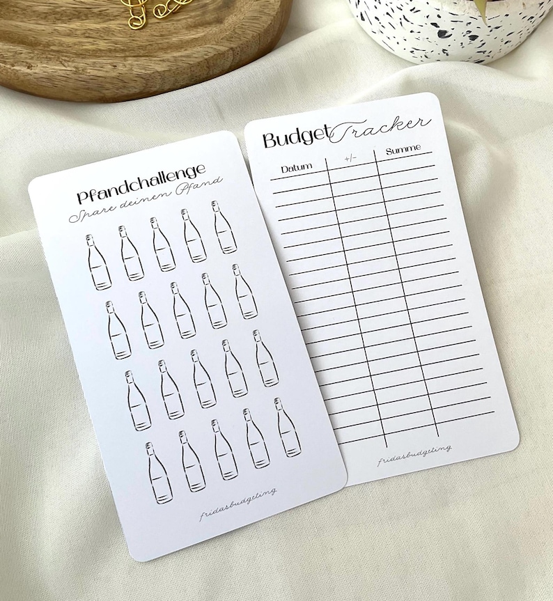 May include: Two white cards with black text and illustrations. One card reads "Pfandchallenge Spare deinen Pfand" with bottle illustrations. The other card says "Budget Tracker" with columns for tracking expenses. Both cards have the text "fridasbudgetling".