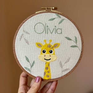 May include: Embroidered hoop art with a yellow and brown giraffe face and green leaves. The name Olivia is embroidered in green thread.