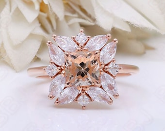 Könnte beinhalten: Ein Verlobungsring aus Roségold mit einem quadratischen Mittelstück, das von kleineren Diamanten umgeben ist. Der Ring hat ein florales Design mit einer Diamanten-Cluster-Umrandung des Mittelstücks.