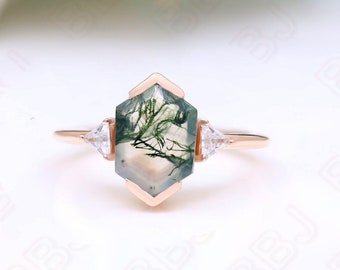 Puede incluir: Un anillo de oro rosa con una piedra hexagonal de ágata verde musgo y dos piedras triangulares transparentes más pequeñas a los lados.