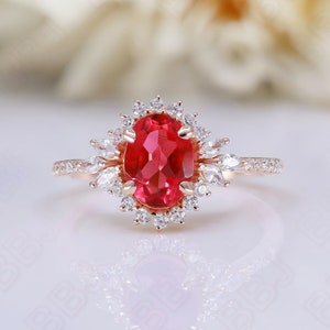 May include: A rose gold engagement ring with a large oval red gemstone surrounded by a halo of smaller white gemstones. The ring has a delicate band with small white gemstones.