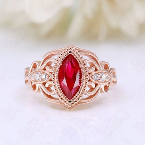 May include: A rose gold ring with a marquise-shaped ruby gemstone surrounded by smaller white gemstones. The ring has a delicate, intricate design.