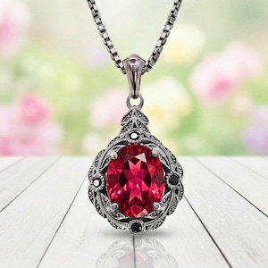 May include: A silver-toned pendant necklace featuring a large, oval-cut, ruby-red gemstone. The pendant is intricately designed with a decorative frame and small black accents. The necklace hangs on a delicate chain.