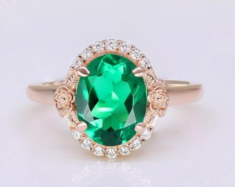 May include: Rose gold ring featuring an oval-cut, vibrant green gemstone. The gemstone is surrounded by a halo of small, clear stones. Floral accents adorn the sides of the ring, adding a touch of elegance to this piece of jewelry.