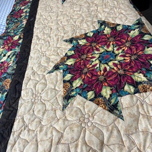 May include: A quilted textile featuring a poinsettia design. The quilt has a beige background with a floral pattern and large, colorful poinsettia shapes in shades of red, green, and gold. A black border frames the design.