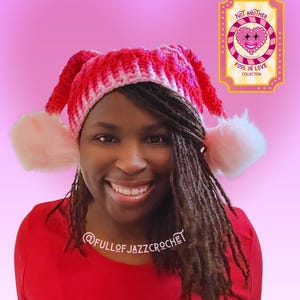Puede incluir: Un gorro de ganchillo hecho a mano en tonos rojos y blancos, con orejeras y pompones rosas. El gorro forma parte de la "Fool in Love Collection" y está etiquetado como "Not Another". También se ve la cuenta @FULLOFJAZZCROCHET.