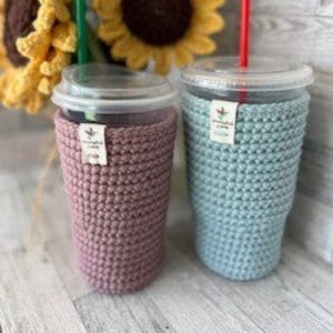 May include: Two crocheted cup cozies in pink and light blue. The cozies are made with a textured stitch and have a small tag attached to each.