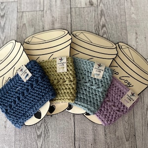Puede incluir: Cuatro calientavasos de punto en diferentes colores: azul, verde, gris y morado. Cada calientavasos tiene una pequeña etiqueta adjunta con el texto "Handmade with Love".
