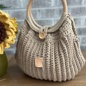 May include: A beige crochet handbag with a wooden handle. The bag features a round wooden handle and a button closure. A small leather tag is attached to the front. The bag is sitting on a wooden surface.