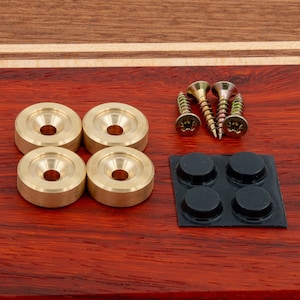 Op de afbeelding: Vier ronde gouden metalen knoppen met een klein gaatje in het midden, drie gouden schroeven en vier zwarte rubberen pads op een vel zwart plastic. De items bevinden zich op een houten oppervlak.