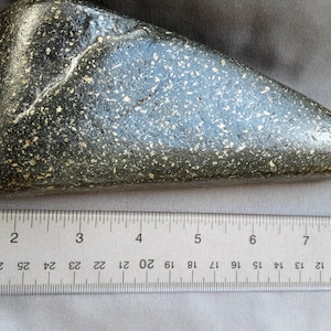 May include: A dark green, speckled stone tool, approximately 8 inches long, with a rounded, pointed end. The tool is measured against a ruler.