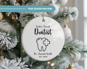 Op de afbeelding: Ronde witte keramische kerstboomversiering met de tekst "Santa's Favorite Dentist" en een tandafbeelding. De versiering bevat ook de naam "Dr. Amanda Smith" en het jaar "EST. 2025". De versiering hangt aan een kerstboom.